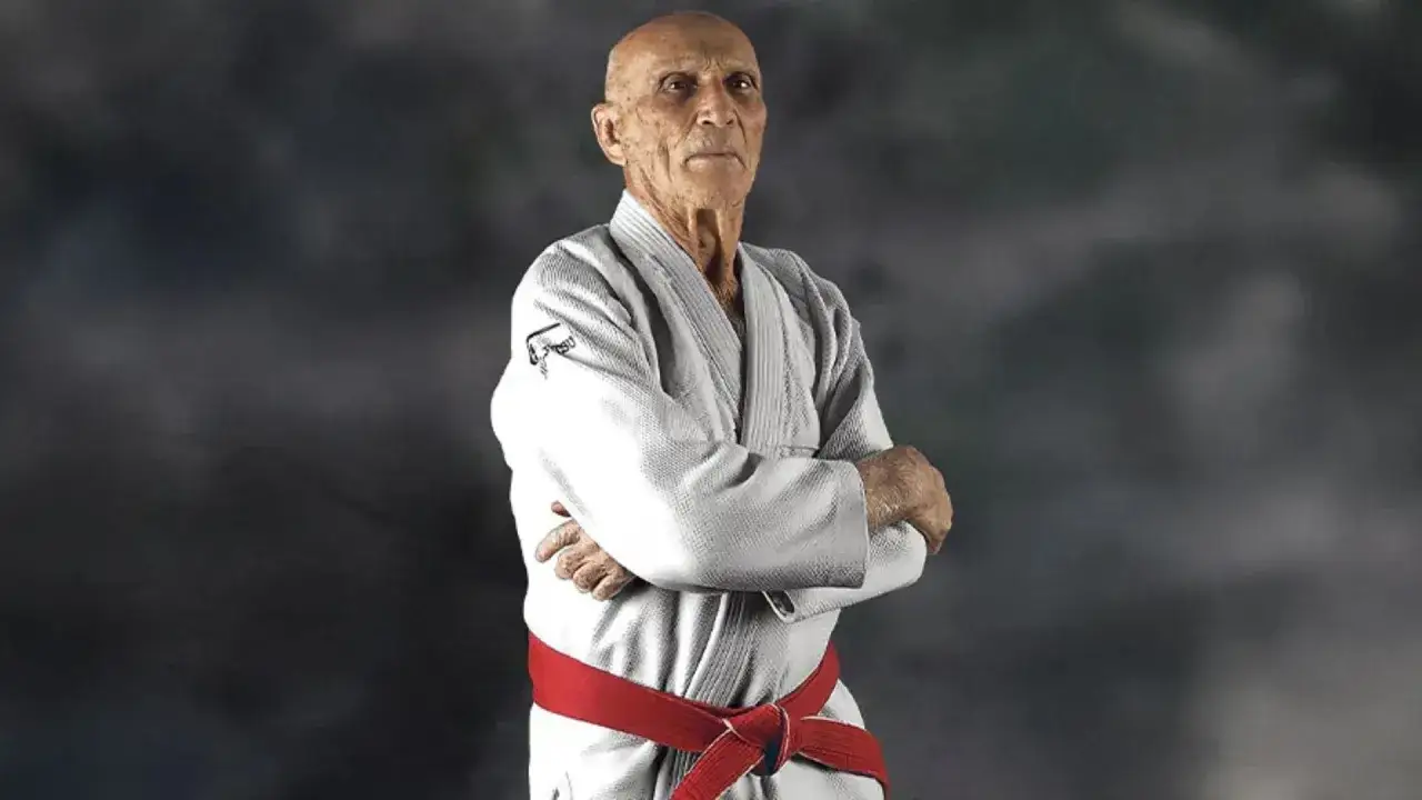Helio Gracie, en &auml;ldre man i vit gi med r&ouml;tt b&auml;lte, st&aring;r med armarna i kors. Hans blick &auml;r fokuserad.