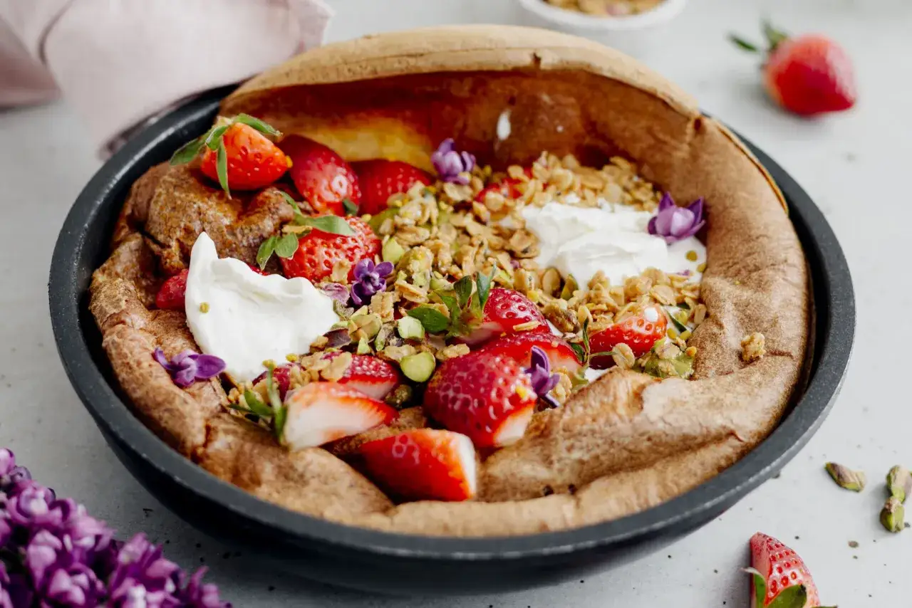 Puszysty dutch baby z truskawkami, granolą i bitą śmietaną, ozdobiony pistacjami i fioletowymi kwiatami.