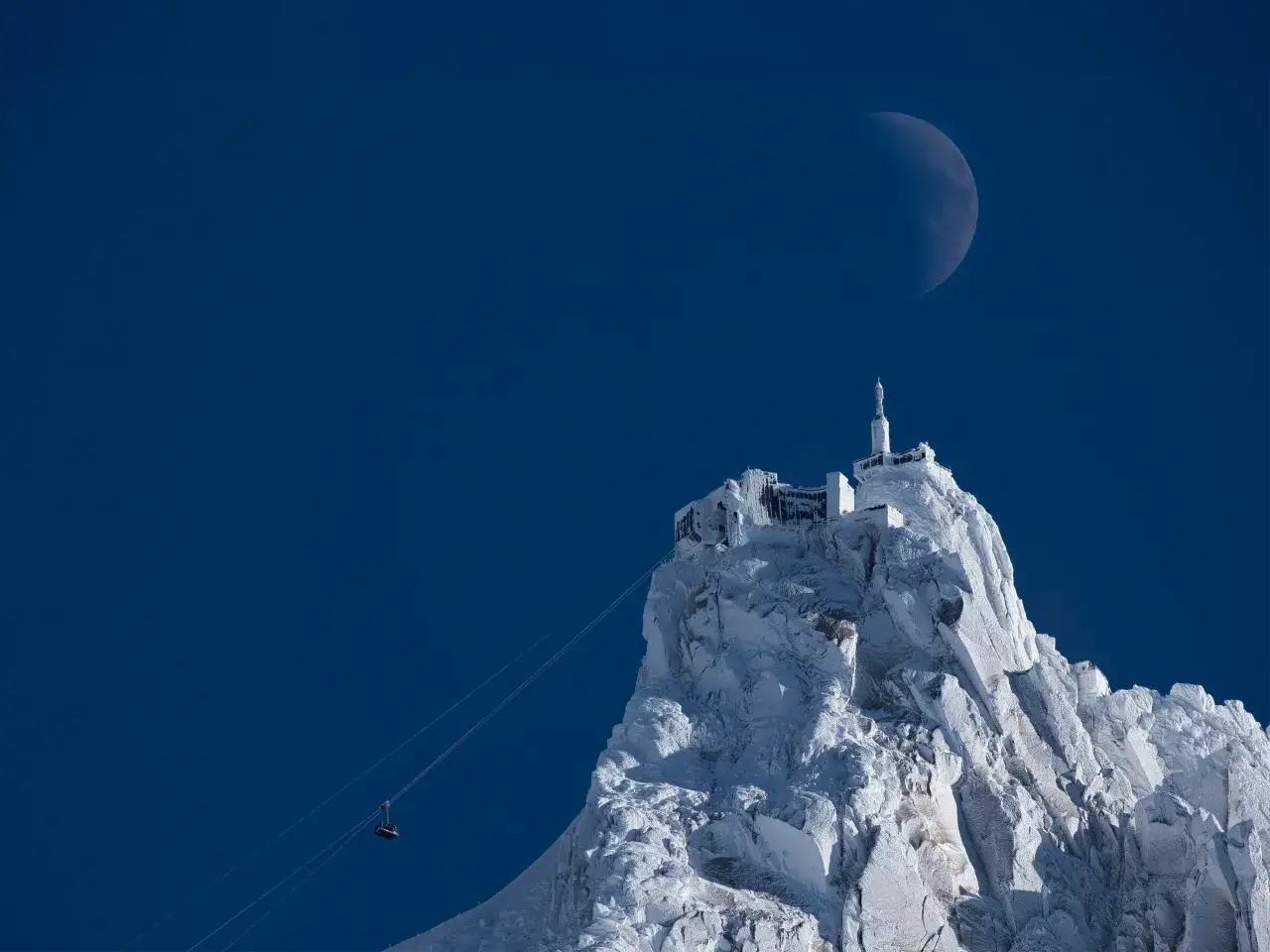Le téléphérique monte vers l'Aiguille du Midi, sommet enneigé sous un ciel bleu profond, avec la lune en croissant visible.