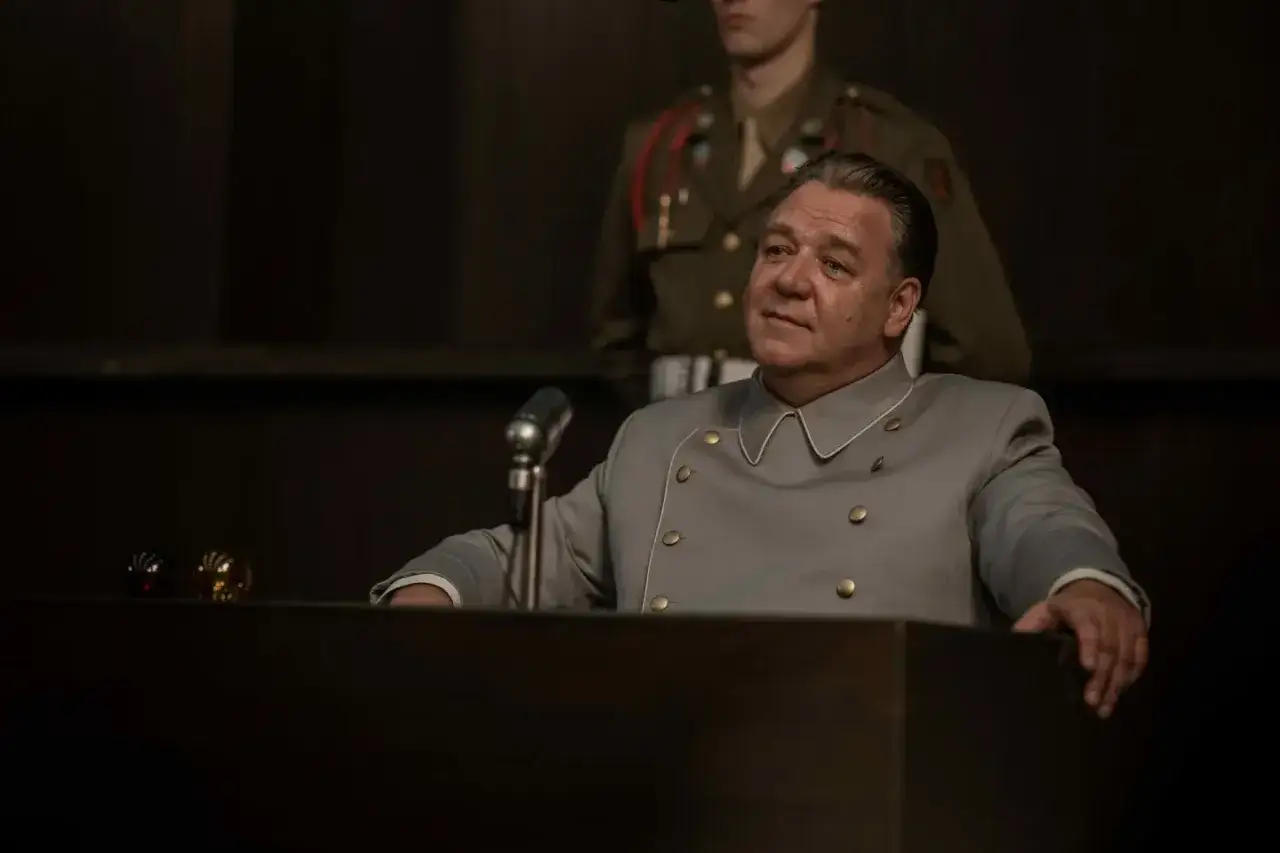 Un uomo in uniforme grigia siede a un podio, con un microfono davanti. Un soldato in uniforme militare &egrave; dietro di lui. La scena evoca un'atmosfera da processo, forse legata a un film su Norimberga.