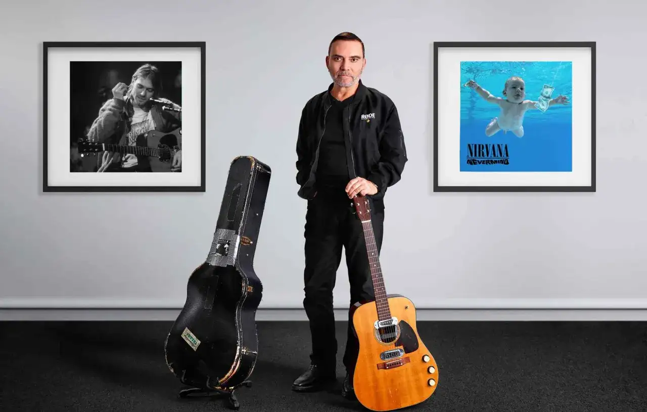 Mężczyzna z gitarą i futerałem, obok obraz&oacute;w z Kurtem Cobainem i okładką 