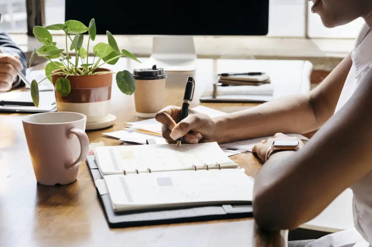 Persona escribe en agenda, con taza y planta de fondo. Un recordatorio: dejar para ma&ntilde;ana lo que puedes hacer hoy.