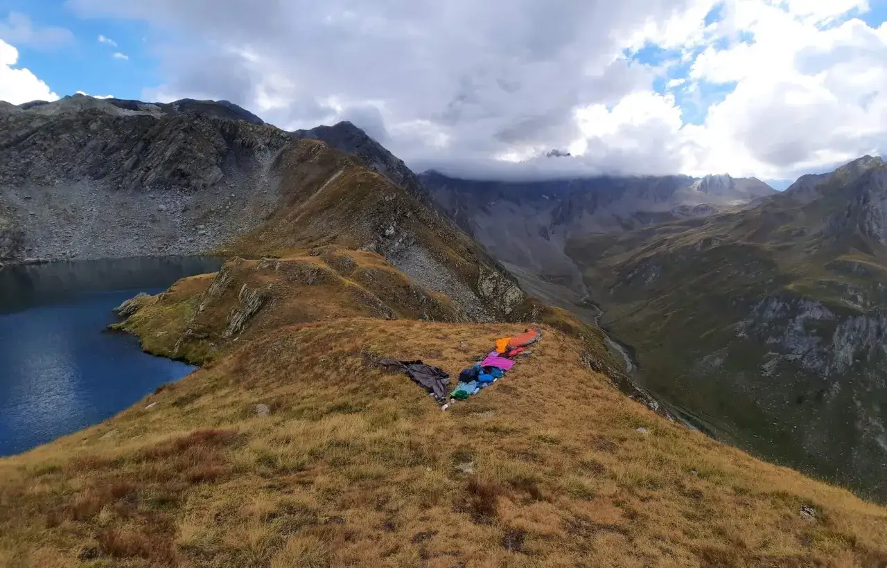 Bunte Schlafsäcke liegen auf einem Bergkamm am via alpina roter weg. Ein See und steile Berge im Hintergrund.