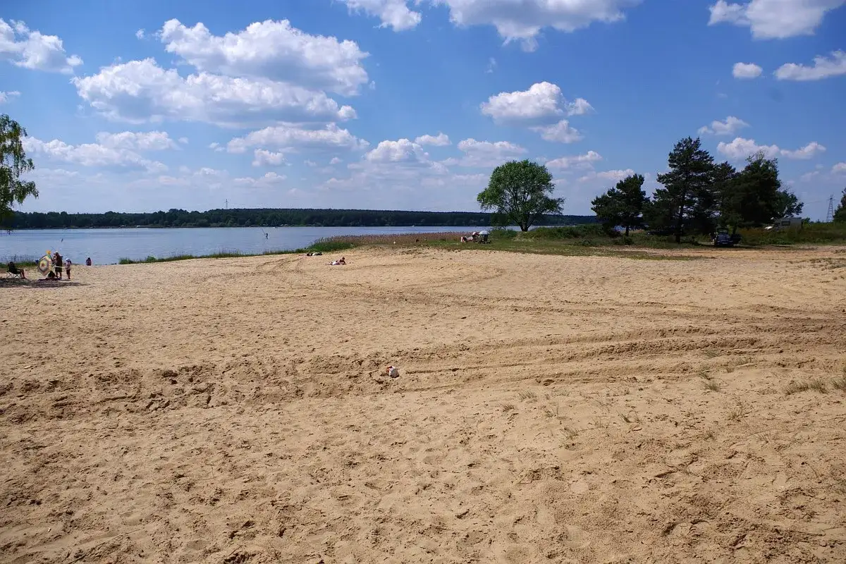 Piękny, piaszczysty **poraj** nad jeziorem. Kilka osób odpoczywa na plaży, ciesząc się słońcem i spokojem.