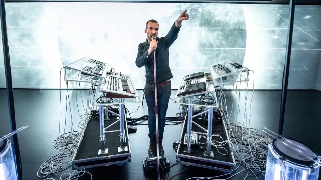 Un homme chante dans un micro, entouré de claviers et de machines, prêt à lancer son groupe electro.