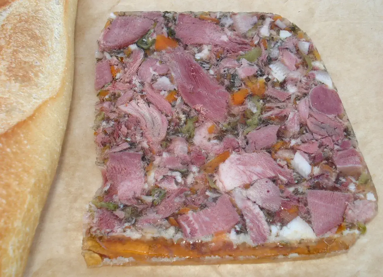 Tranche de fromage de t&ecirc;te avec morceaux de viande, l&eacute;gumes et gel&eacute;e, &agrave; c&ocirc;t&eacute; d'une baguette de pain.