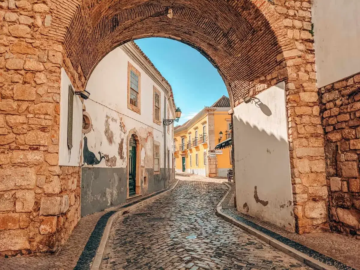 Gepflasterte Gasse in der Faro Portugal Altstadt unter einem alten Ziegelsteinbogen. Sonnige Häuser säumen den Weg.