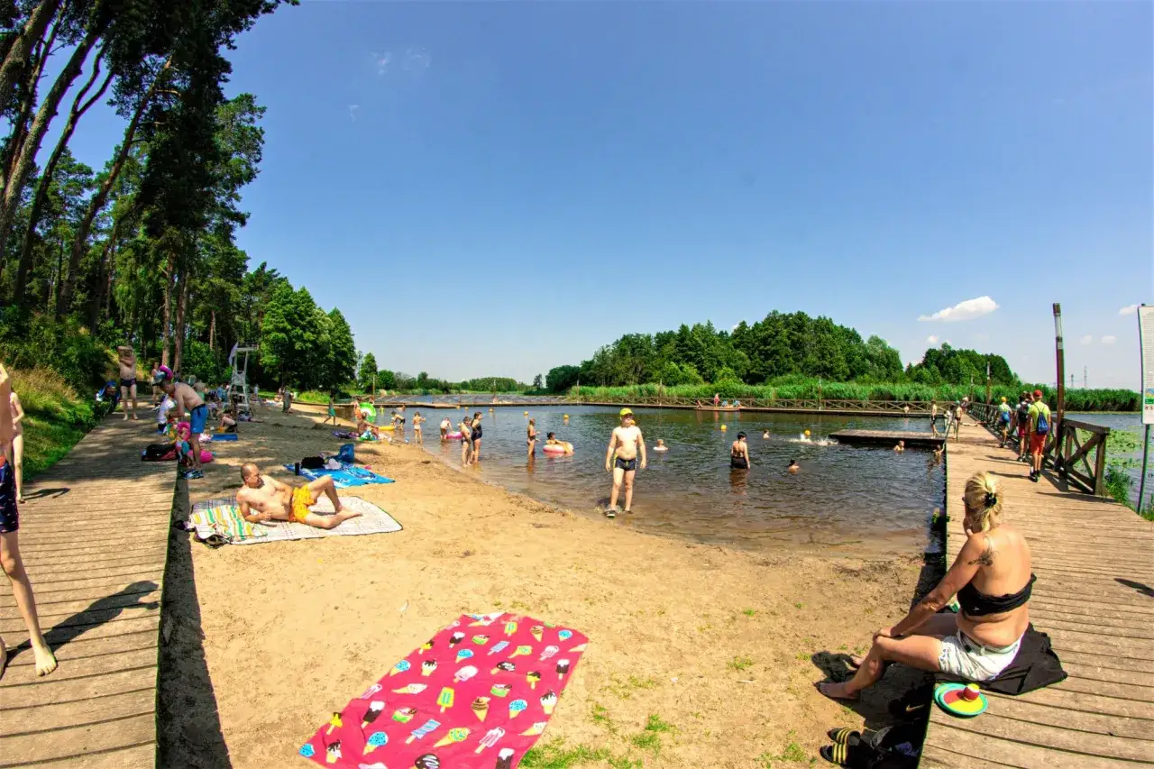 Ludzie odpoczywają i kąpią się nad **jeziorem kozienickim**. Słoneczny dzień sprzyja plażowaniu i wodnym zabawom.