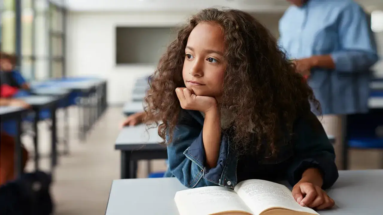 Flicka med lockigt hår sitter vid ett skrivbord med en öppen bok. Hon ser ut att vara djupt försjunken i tankar, kanske kämpar med koncentrationen som vid barn depression eller adhd.