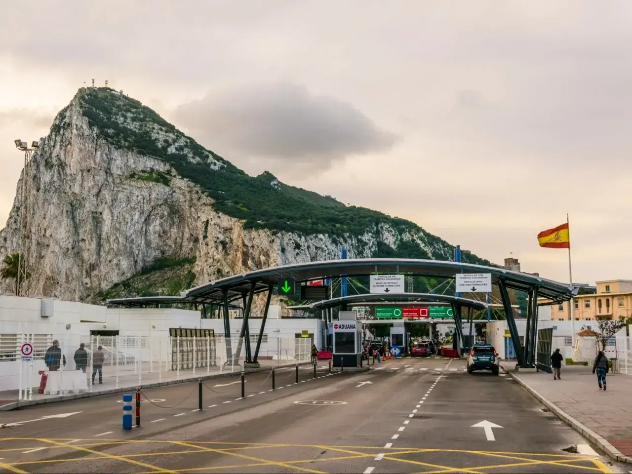 Wjazd do Gibraltaru pod skalistym Gibraltarem. Na granicy widać budynki celne i flagę Hiszpanii.