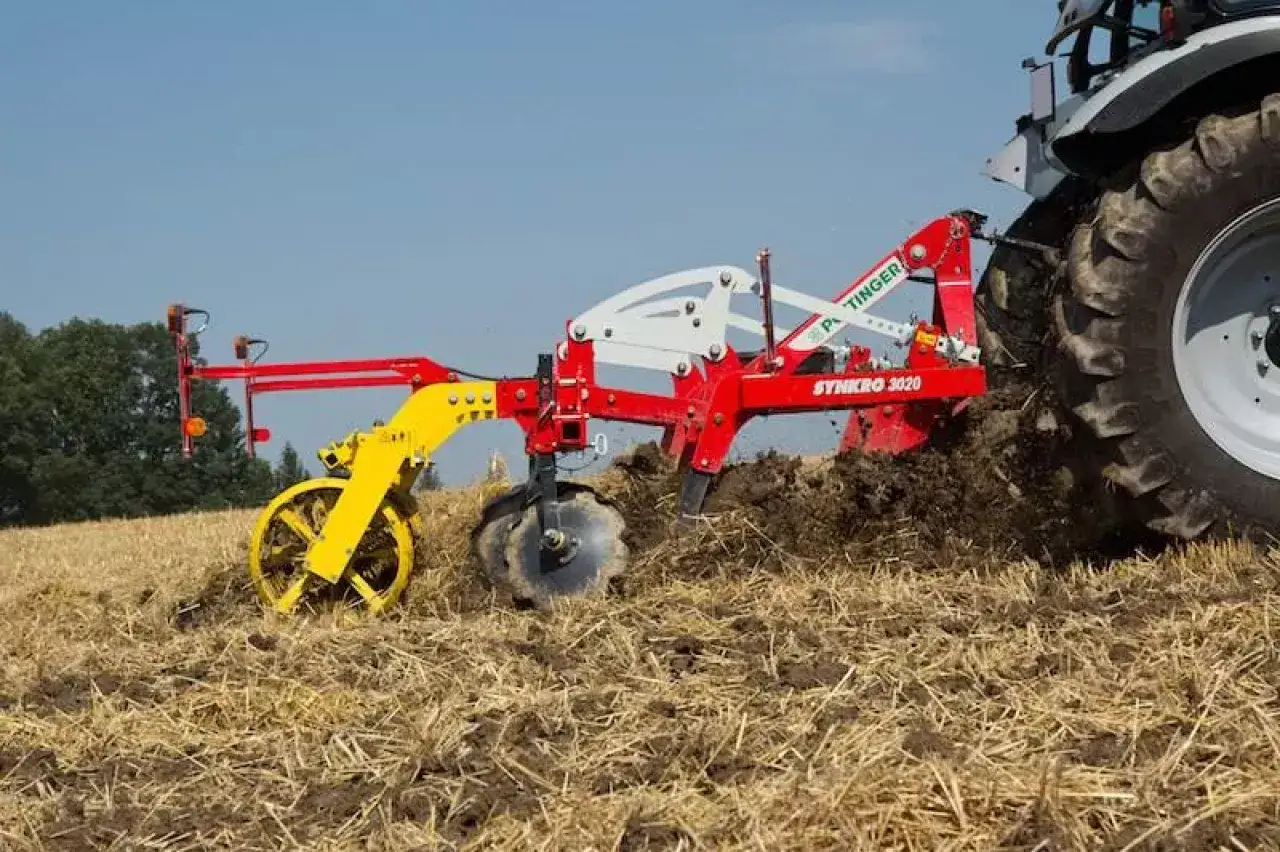 Czerwony kultywator P&ouml;ttinger Synkro 3020 pracuje na polu, spulchniając ziemię. To maszyna rolnicza do uprawy gleby.