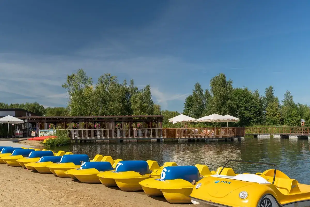 Żółte rowerki wodne czekają na plaży nad zalewem żyrardowskim. W tle drewniana kładka i zielone drzewa.