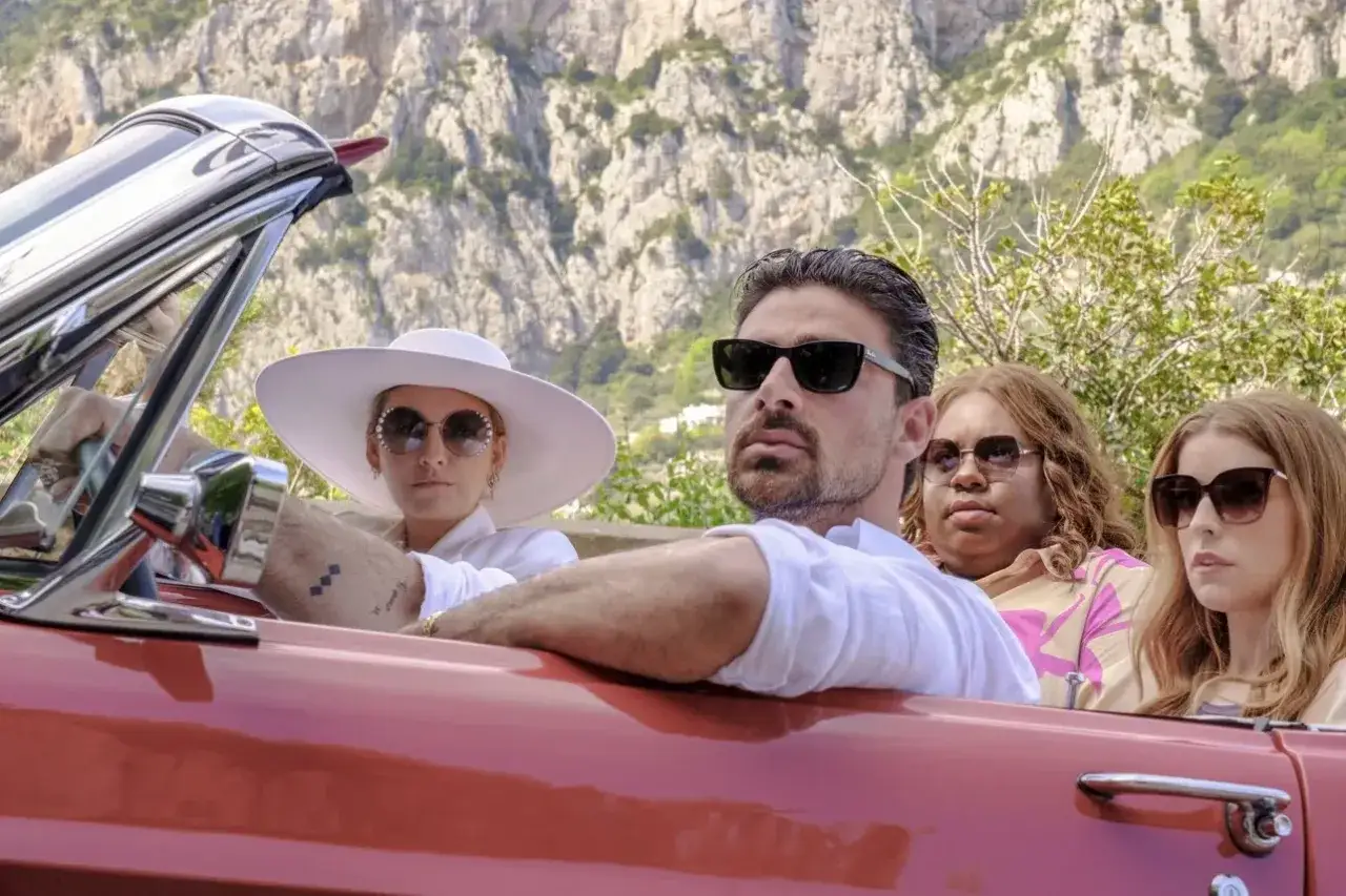 Un uomo e tre donne in un'auto d'epoca rossa, con un paesaggio montuoso sullo sfondo. Un semplice favore per un viaggio indimenticabile.