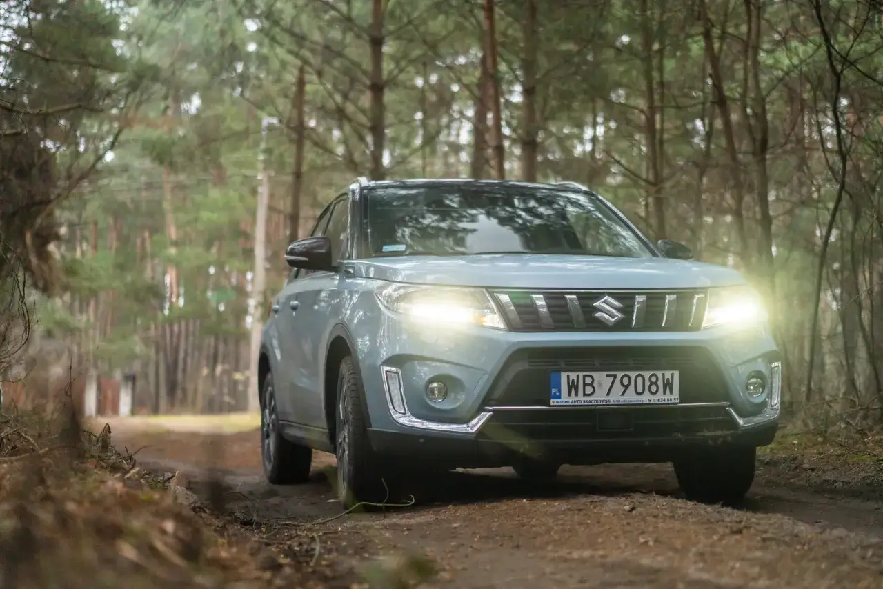 Jasnoszary Suzuki Vitara Hybrid na leśnej drodze. Reflektory świecą, a auto wygląda gotowe do przygody.