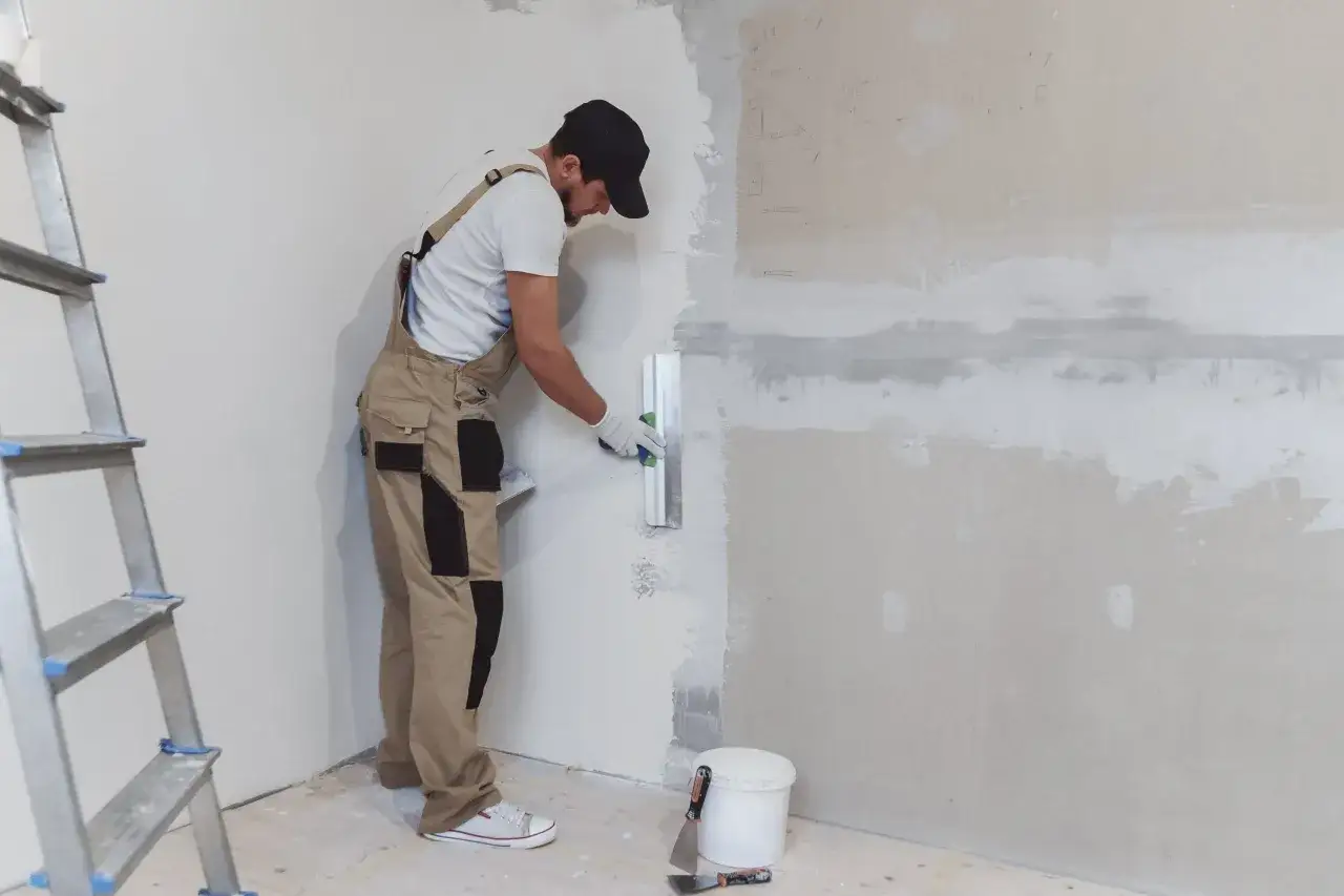 Obrero aplica yeso en pared. Pregunta: cuanto tarda el yeso en secar para poder pintar. Escalera y herramientas a la vista.