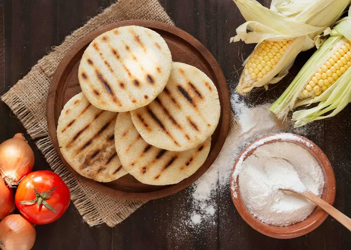 Arepas colombianas recién hechas, doradas y con marcas de parrilla, junto a ingredientes frescos como maíz, tomate y cebolla.