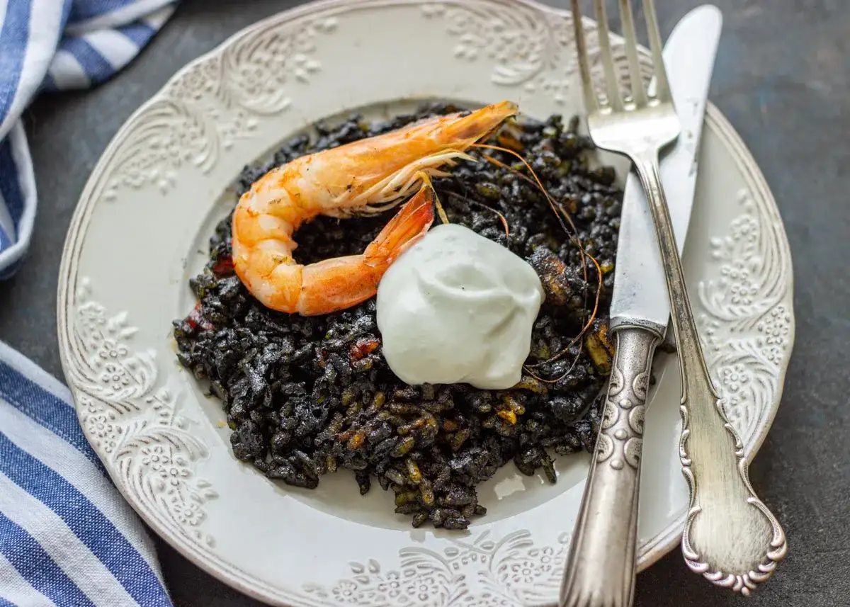 Un plato de arroz negro con un langostino y una quenelle de crema. Un tenedor y un cuchillo descansan a la derecha.