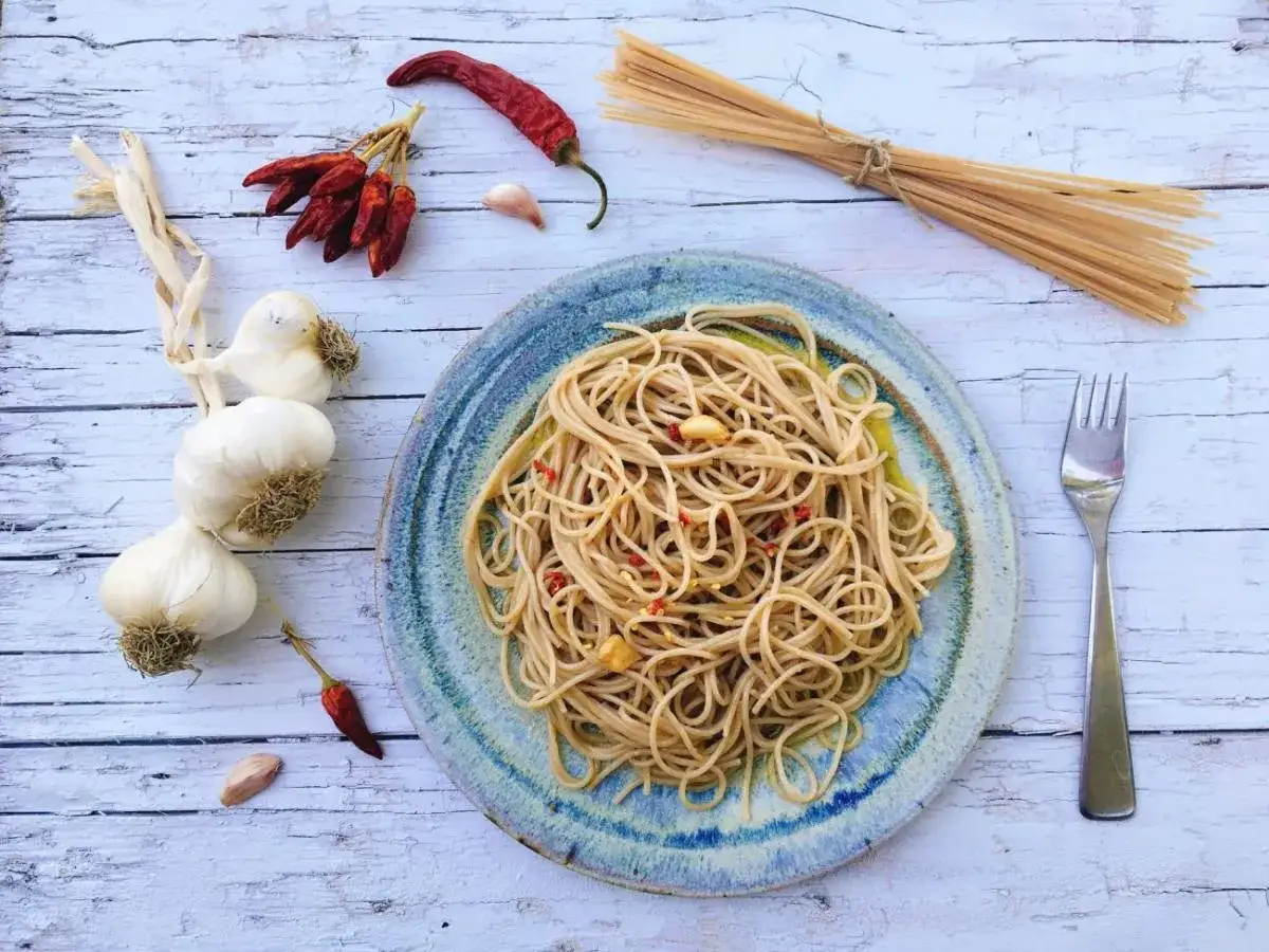 Espaguetis al ajillo servidos en un plato r&uacute;stico, acompa&ntilde;ados de ajos, guindillas secas y un manojo de pasta cruda.