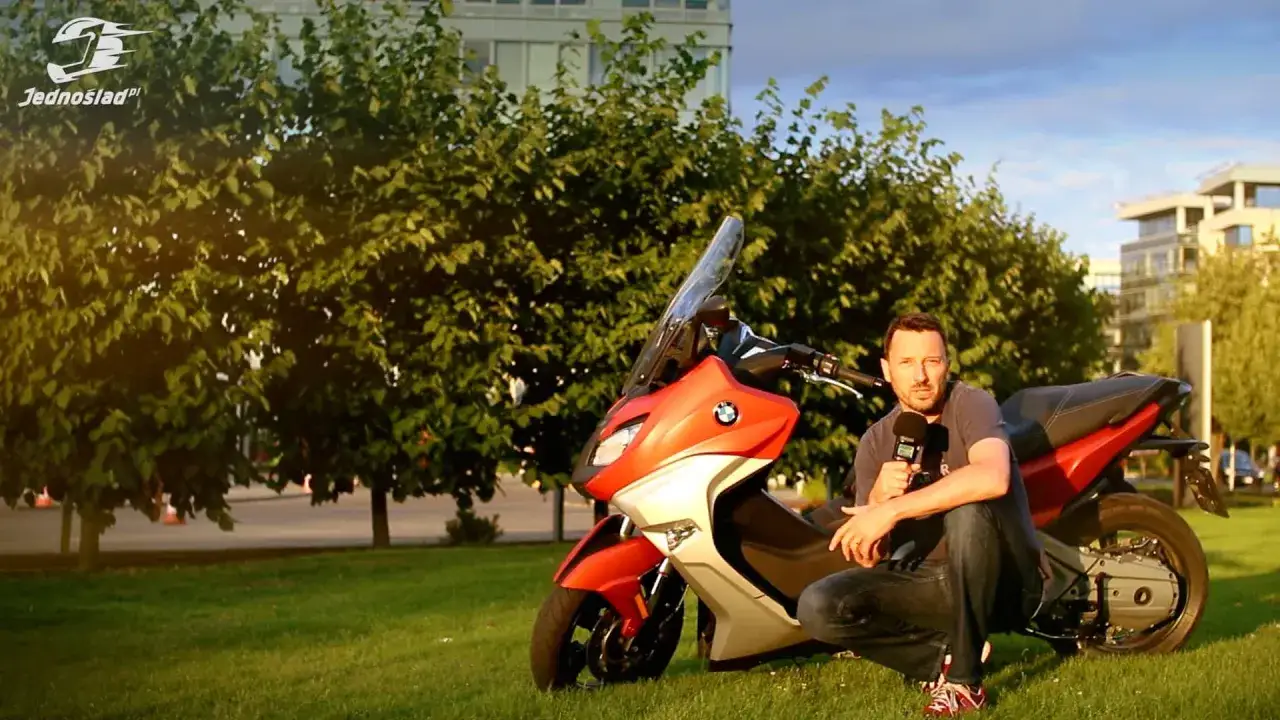 Mężczyzna z mikrofonem przy pomarańczowo-białym skuterze BMW C650 Sport na tle drzew i budynków.