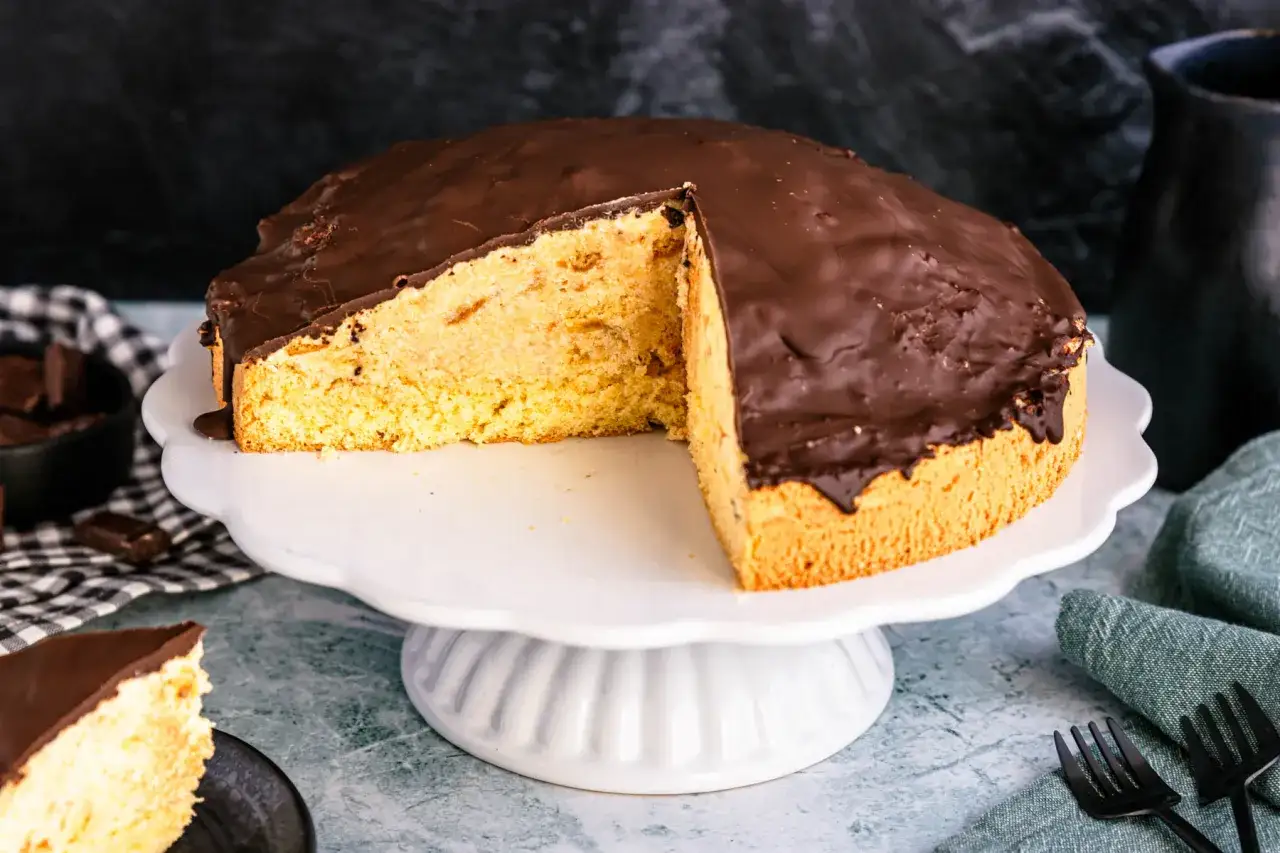Ein saftiger Schokoladenkuchen, der nach einem einfachen rumbombe rezept gemacht wurde, steht auf einem weißen Tortenständer. Ein Stück fehlt.