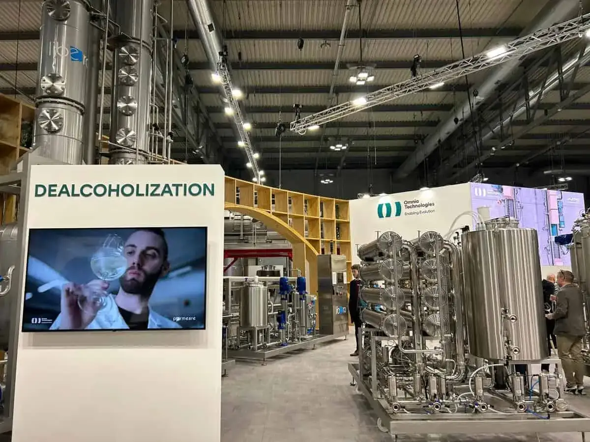 Stand fieristico con macchinari per la dealcolizzazione. Un video mostra un uomo che osserva un bicchiere di vino.