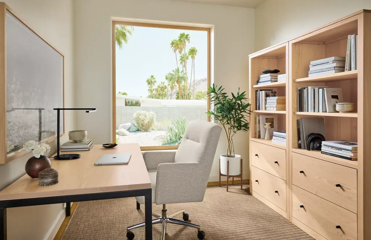 A bright, modern home office with a light wood desk, a comfortable chair, and a tall bookshelf. This space offers great personal office decor ideas, featuring a large window with a desert view.