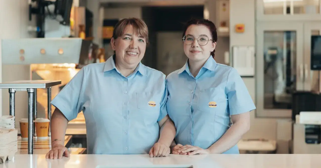 Dwie uśmiechnięte pracownice McDonald's w jasnoniebieskich koszulach z logo.