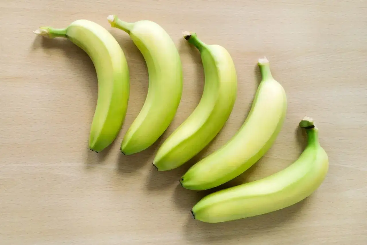 Pięć zielonych bananów ułożonych na drewnianym blacie. Czy banany są zdrowe na jelita? Tak, są bogate w błonnik.