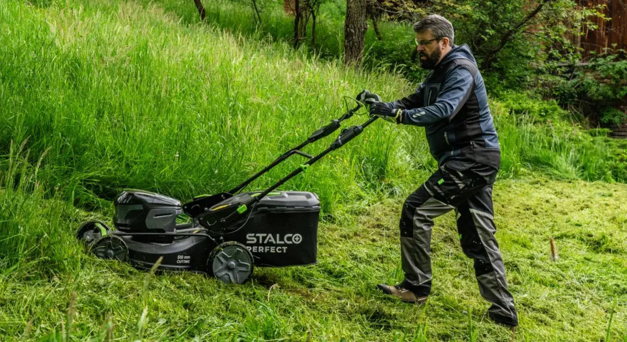 Mężczyzna z kosiarką Stalco Perfect w wysokiej trawie. Zastanawia się, na jaką wysokość kosić trawę, by uzyskać idealny efekt.