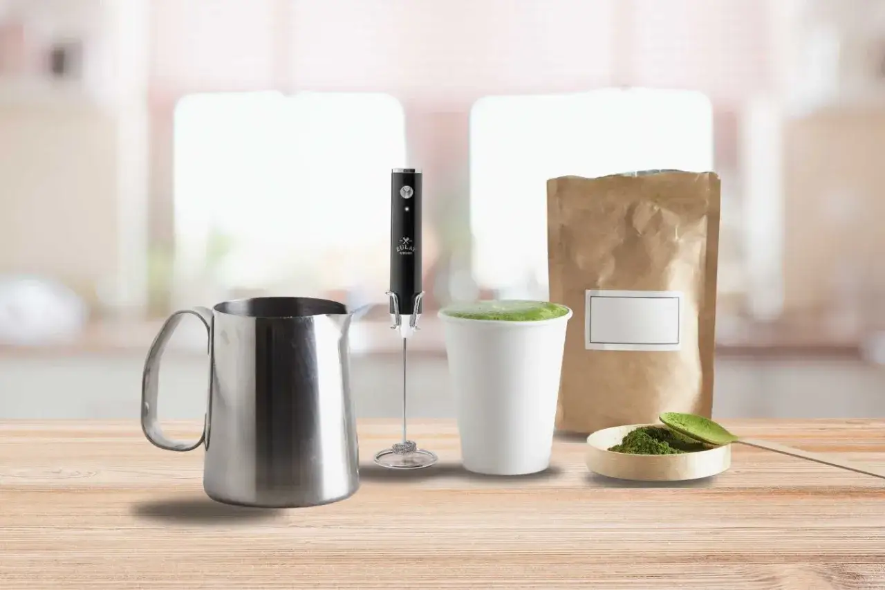 Perfekter Matcha ohne Besen: Milchkanne, elektrischer Aufsch&auml;umer, Tasse mit cremigem Matcha und Pulver auf einem Holztisch.