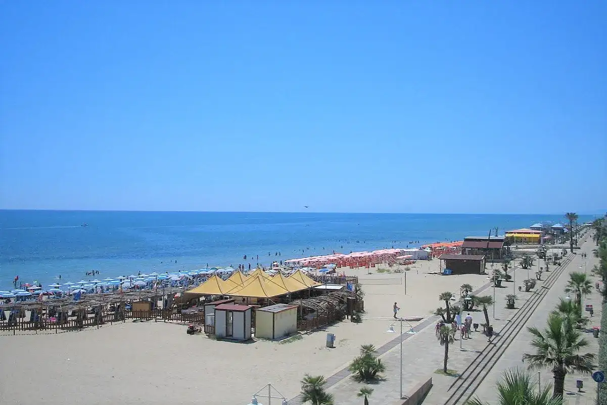 Una giornata di sole a Metaponto Lido. Spiaggia affollata con ombrelloni colorati, mare azzurro e una passeggiata alberata.