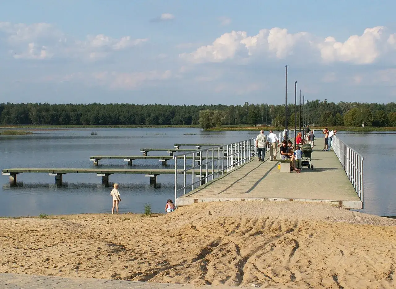 Ludzie spacerują po pomoście nad zalewem Borki. W tle las, na piasku bawią się dzieci.