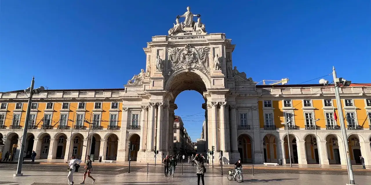 En solig dag i Lissabon gamla stan. En imponerande triumfb&aring;ge dominerar torget, med folk som promenerar och cyklar.