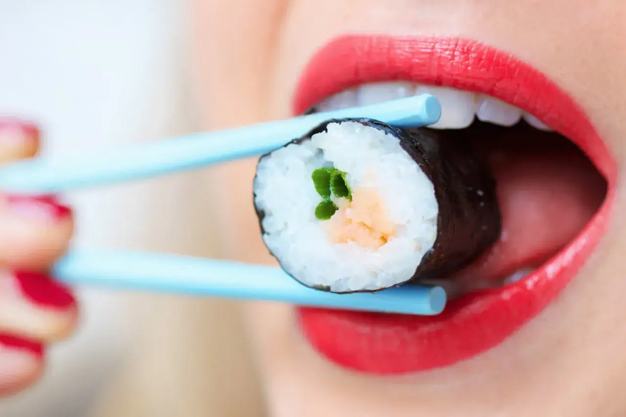 Usta z czerwonymi ustami biorą pałeczkami kawałek sushi. Idealny spos&oacute;b, jak jeść sushi.