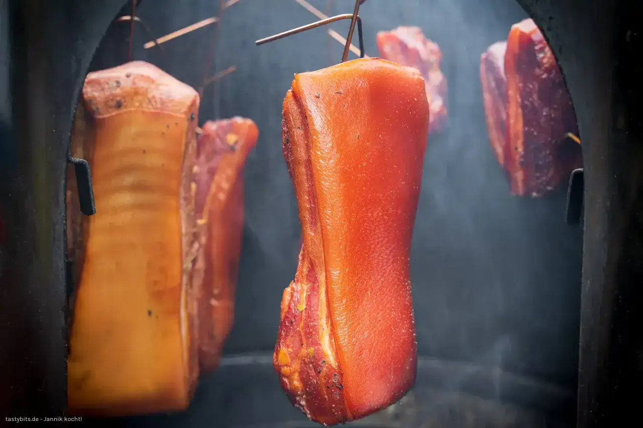 Fleischst&uuml;cke h&auml;ngen im Rauch, bereit f&uuml;r das **bacon selber machen**. Der Prozess des R&auml;ucherns verleiht dem Speck seine charakteristische Farbe und Aroma.