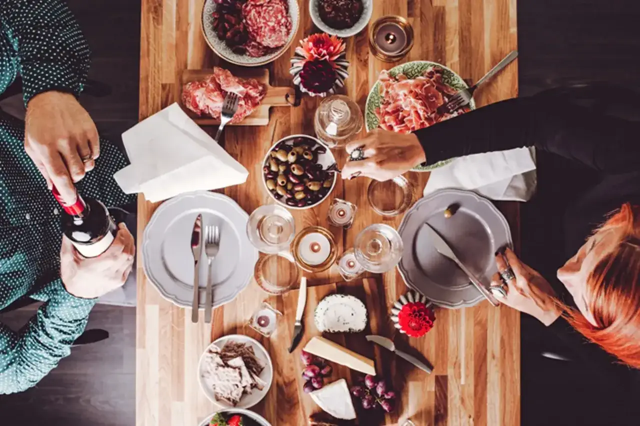 Un homme ouvre une bouteille de vin pour un d&eacute;jeuner convivial. Sur la table en bois, charcuterie, olives, fromages, raisins et crackers invitent au partage.