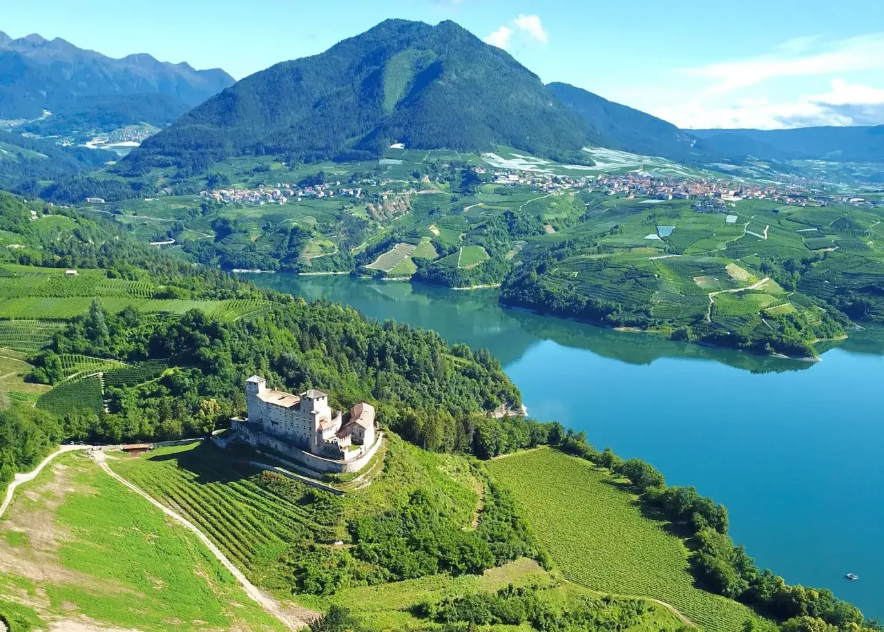 Castello medievale su una collina verdeggiante, circondato da vigneti e affacciato su un lago azzurro nel Val di Non.