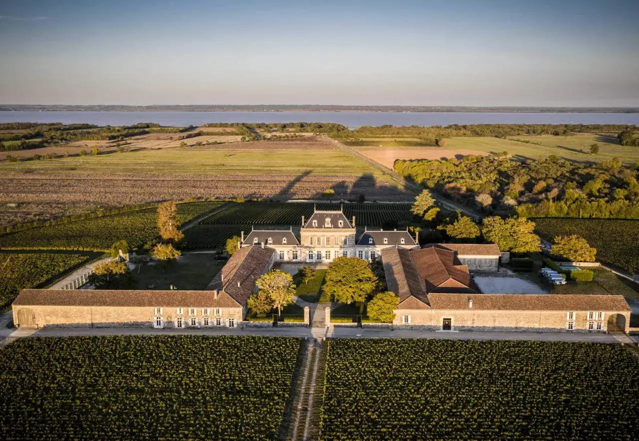 Vue aérienne du **Château Pey La Tour** baigné par le soleil couchant, entouré de vignobles à perte de vue et d'une étendue d'eau.