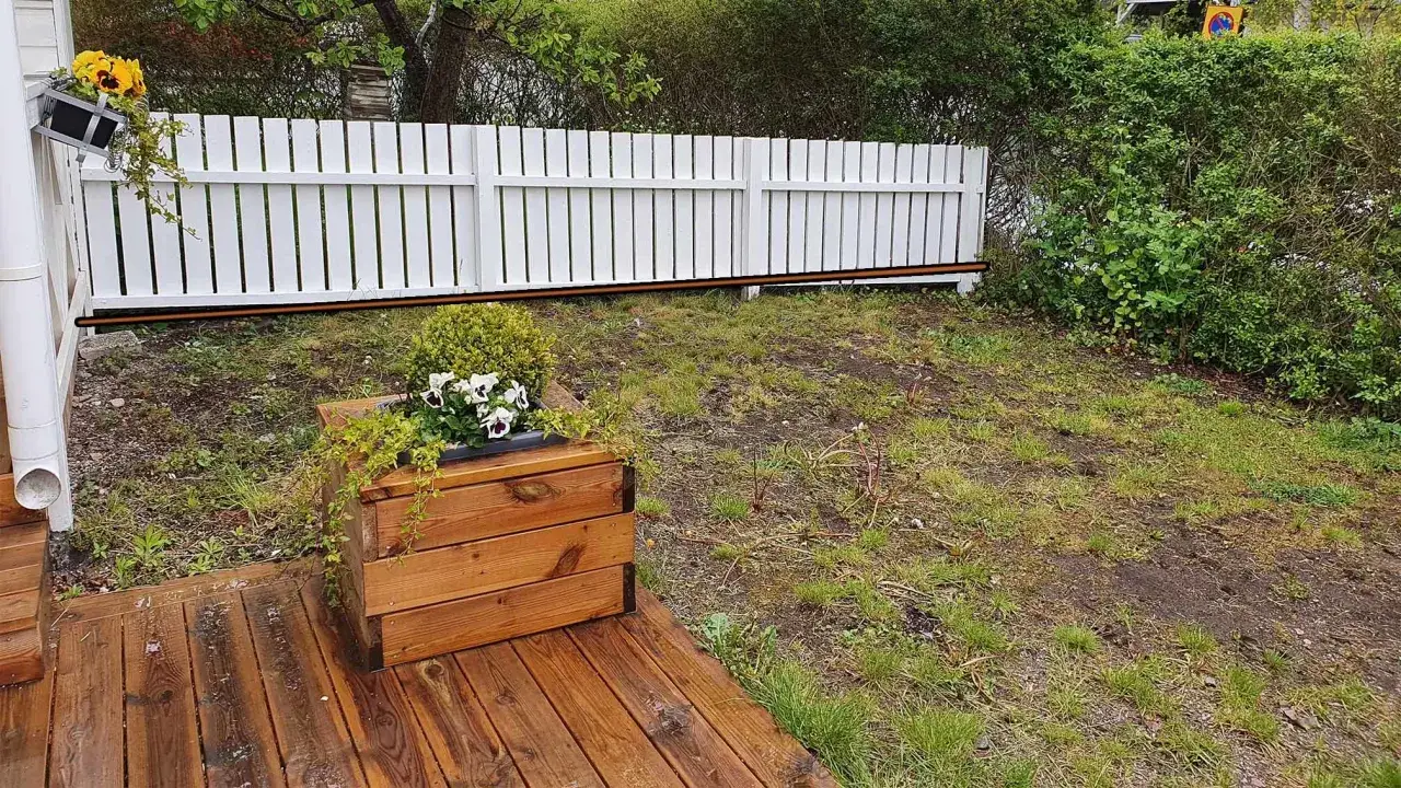 Trädäck med en **altan nivåskillnad** mot en sluttande gräsmatta. En vit plankstaket avgränsar.