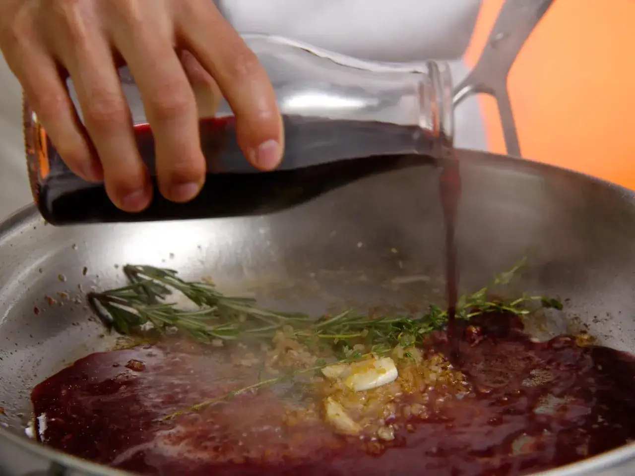 Une main verse un liquide rouge fonc&eacute; dans une po&ecirc;le contenant de l'ail, des herbes et du jus de viande.