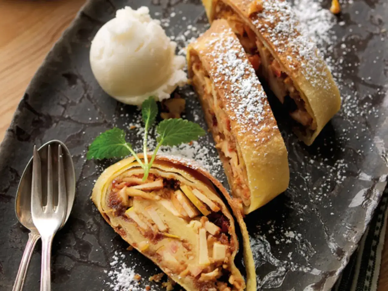 Ein Stück Apfelstrudel, bestäubt mit Puderzucker, neben einer Kugel Vanilleeis und einem Löffel mit Gabel.