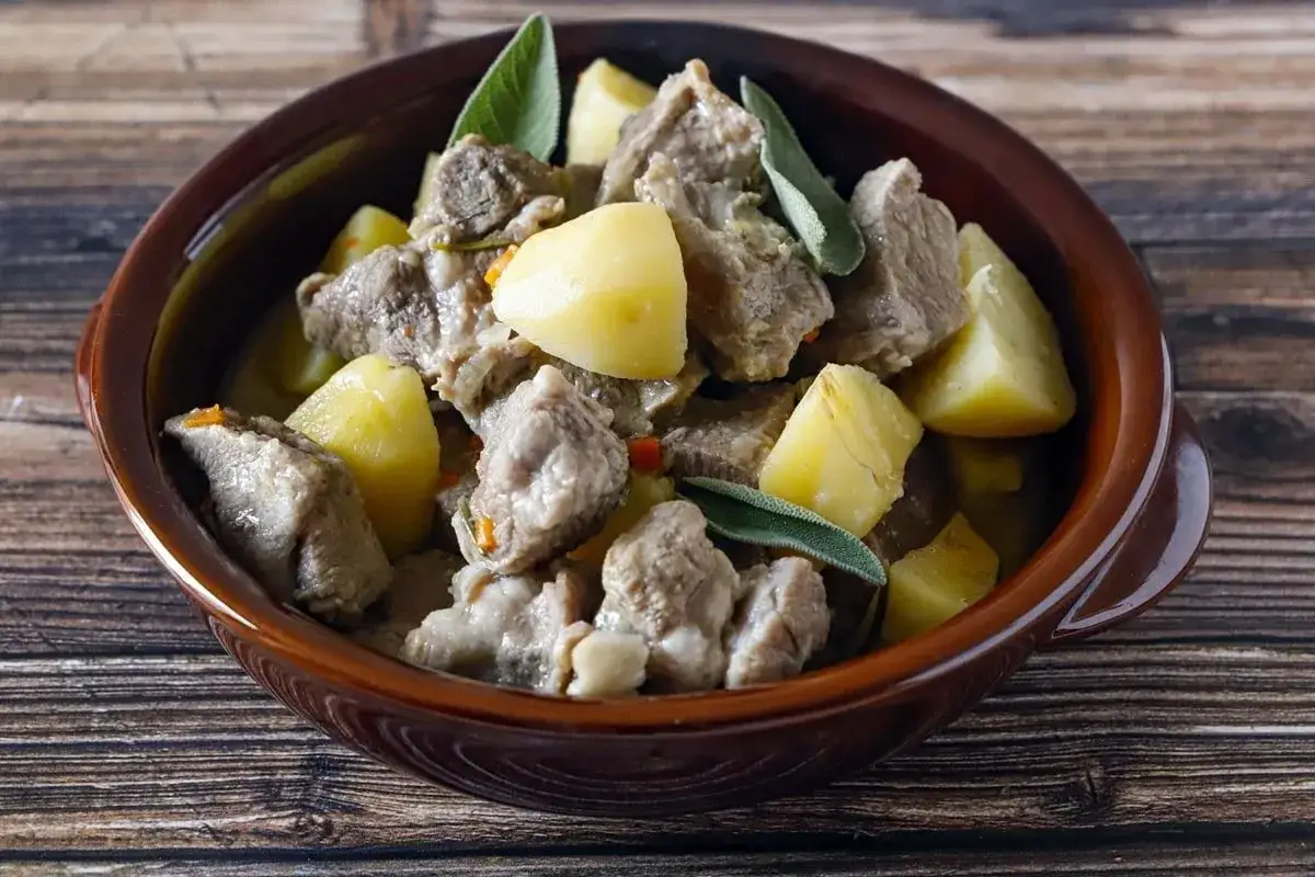Un saporito spezzatino in bianco con patate e salvia, servito in una ciotola di terracotta su un tavolo di legno.