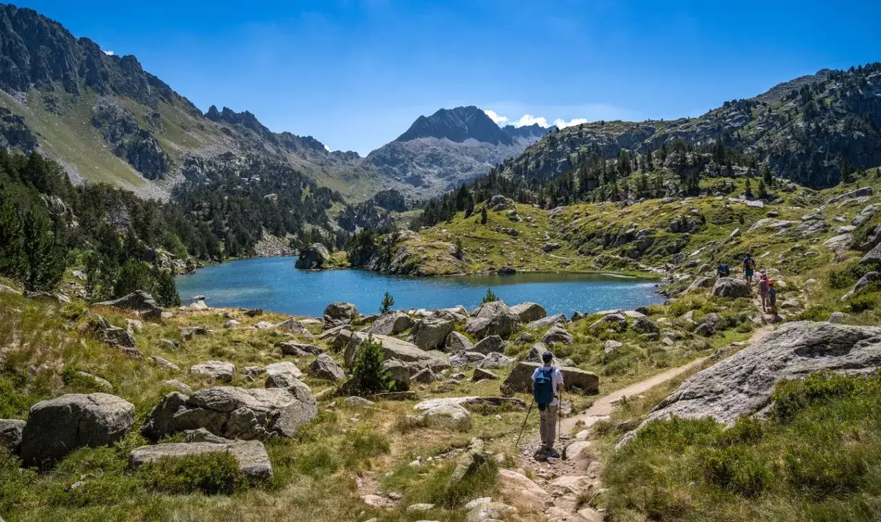 Senderistas disfrutan de un día soleado en el **Valle de Arán**, rodeados de montañas, un lago azul y rocas.