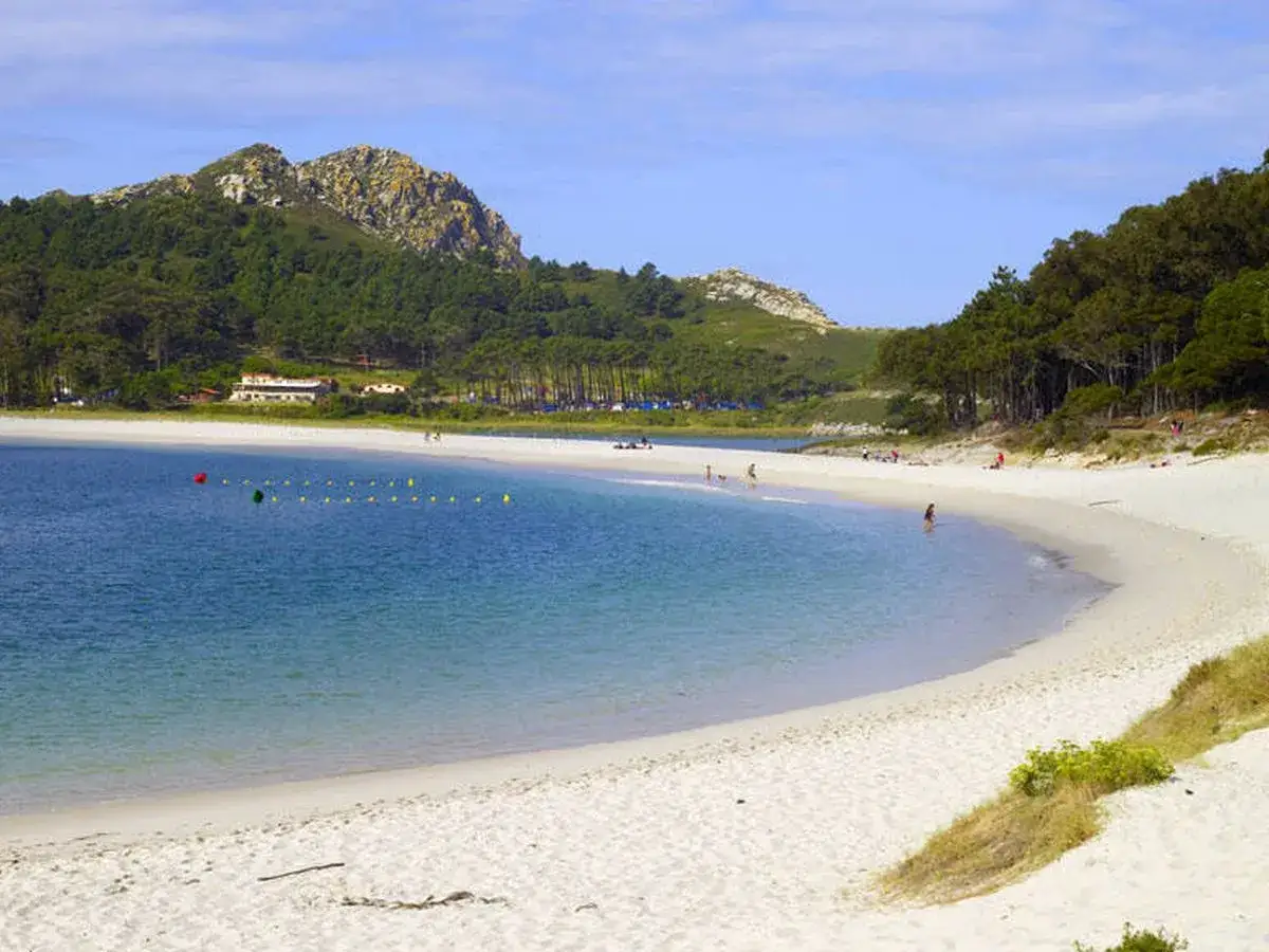 La **playa de Arealonga** se extiende con arena blanca y aguas turquesas, rodeada de pinos y montañas. Pocas personas disfrutan del sol y el mar.