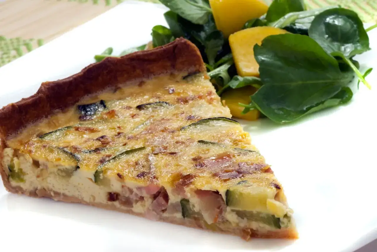 Rebanada de quiche de calabacín y bacon, con trozos de queso y una ensalada verde al lado.