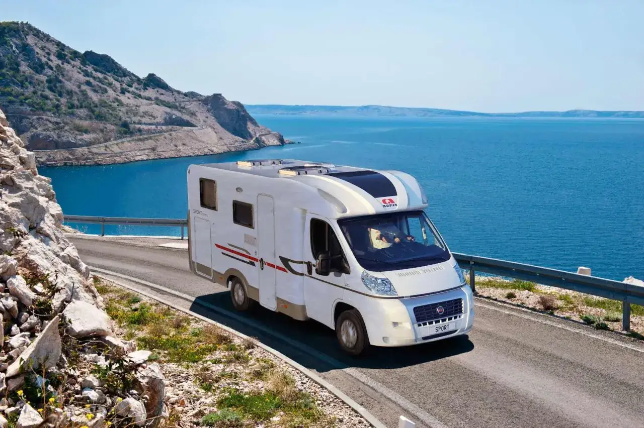 Un camping-car Adria roule sur une route côtière en Sardaigne, avec la mer bleue et des montagnes rocheuses en arrière-plan.