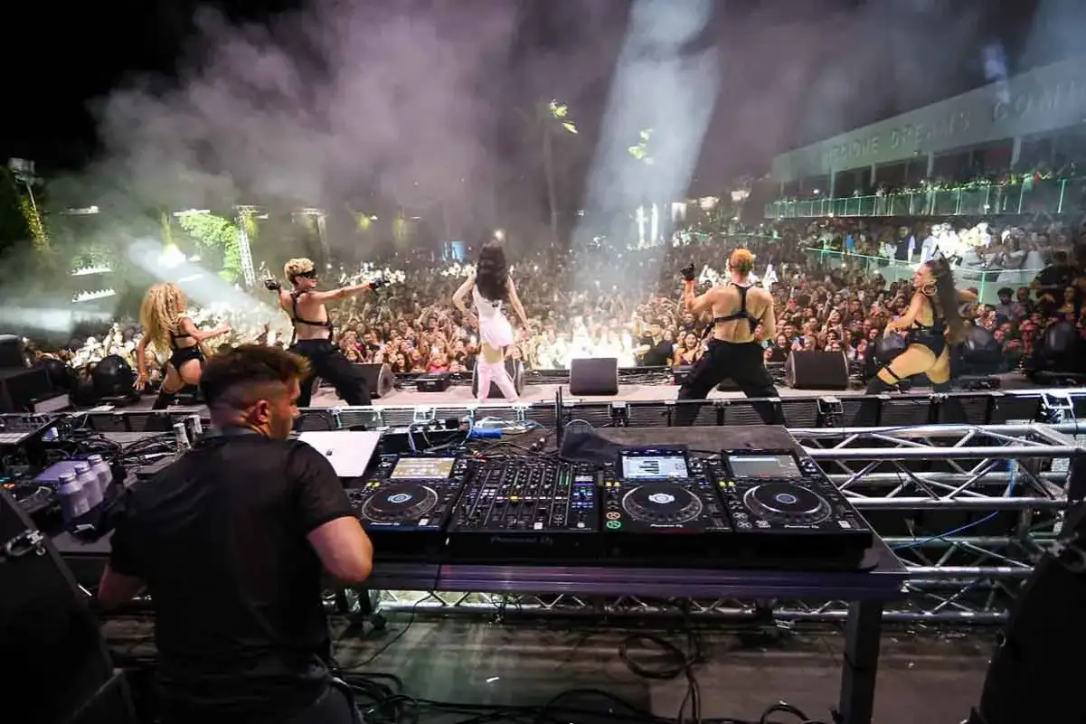 Anna cantante sul palco con ballerini e DJ, davanti a una folla entusiasta.