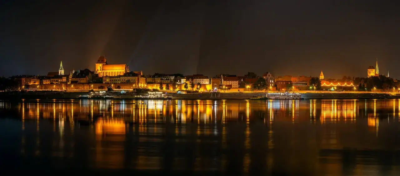 Nocny bulwar filadelfijski w Toruniu, oświetlony ciepłym blaskiem latarni i budynków, odbija się w spokojnej tafli Wisły.