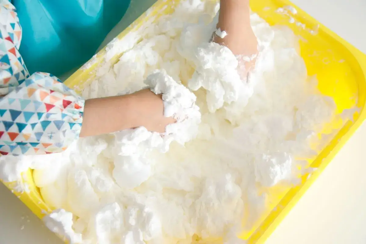 Manos de niño jugando con slime de espuma de afeitar en una bandeja amarilla.