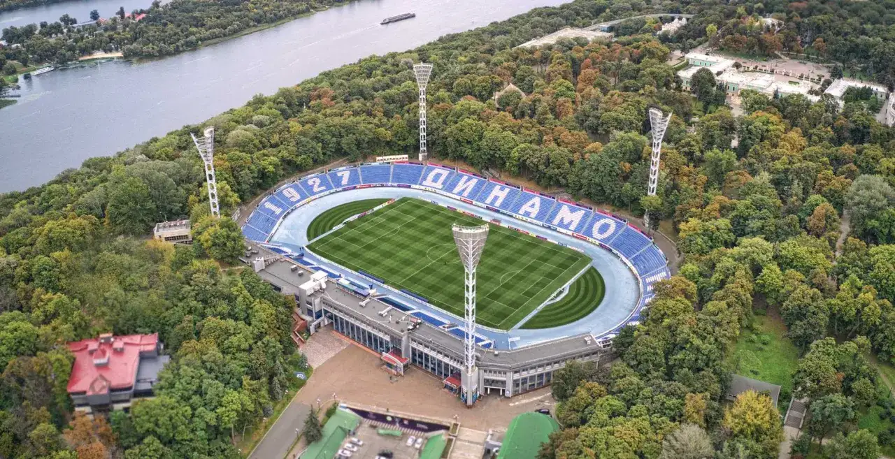 Stadion Dynamo Kijów otoczony zielenią, z boiskiem i niebieskimi trybunami. W tle rzeka i budynki.